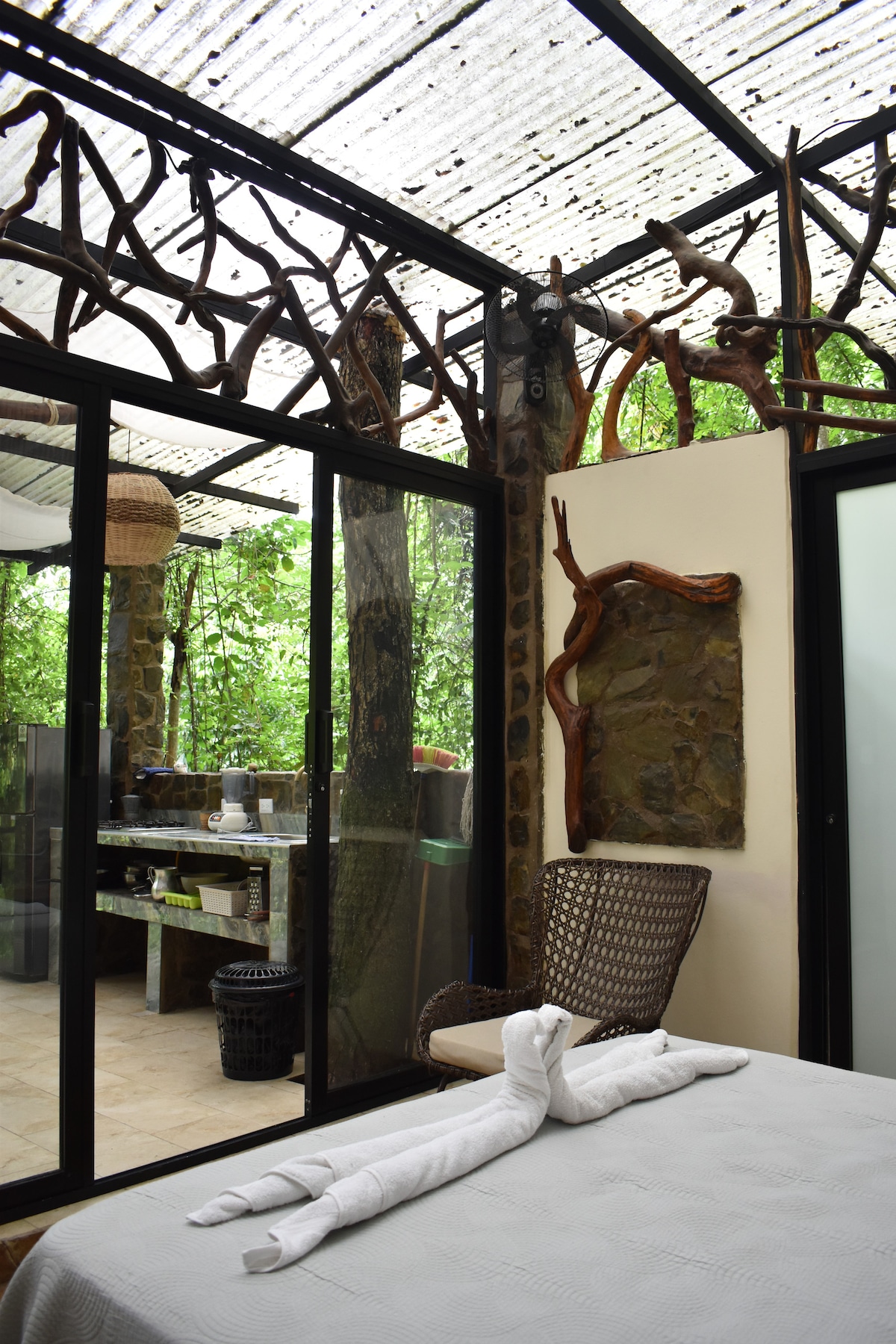 The image captures a serene interior space featuring large glass doors that open to an outdoor kitchen area. Natural wood accents adorn the ceiling, creating a connection to the surrounding greenery. A neatly made bed with a decorative towel arrangement sits in the foreground.
