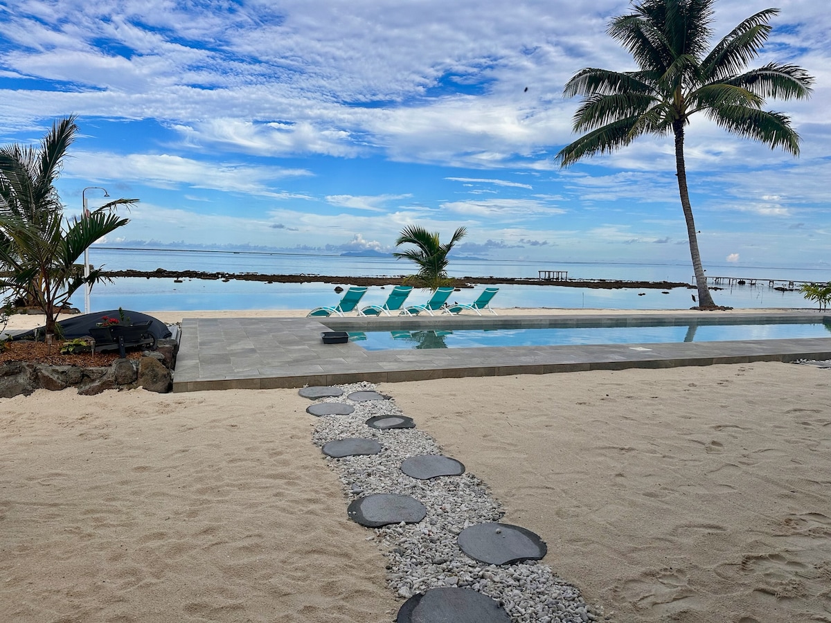 A serene beachfront view features a clear pool with lounge chairs positioned around it. Palm trees provide shade, while the ocean stretches into the distance under a partially clouded sky. Smooth stone pathways lead through the sandy area, enhancing the tranquil atmosphere.