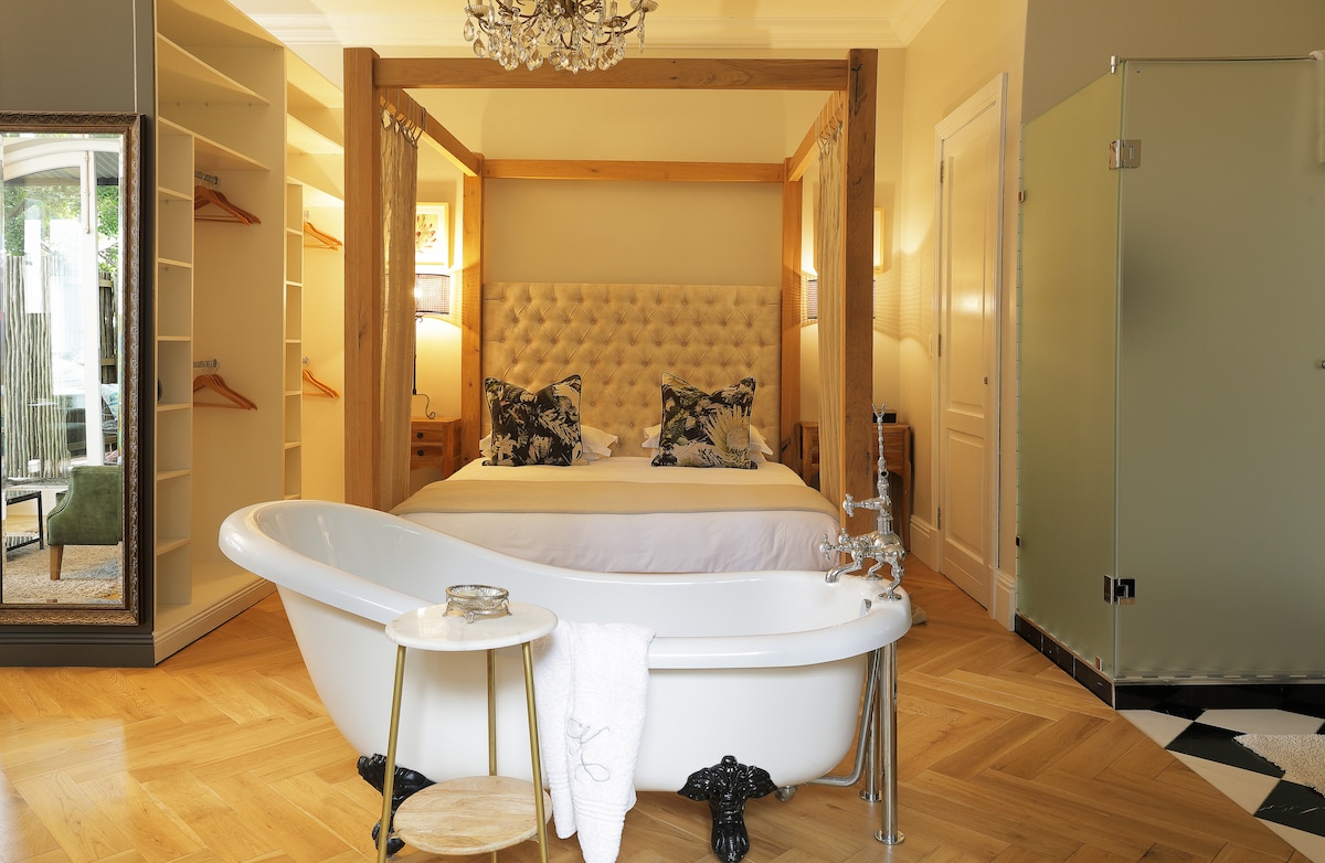 A spacious bedroom features a tufted bed framed by elegant wooden posts. A vintage freestanding bathtub is positioned in the foreground, accompanied by a small wooden table. Shelving units with hanging space line one side, while natural light enters through a nearby glass door.