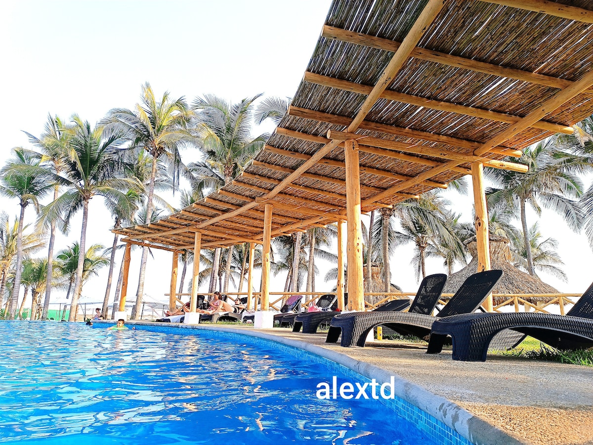 A palm-lined area features a spacious common pool, with lounge chairs positioned beneath a thatched-roof structure providing shade. The pool's crystal-clear water reflects the surrounding greenery, creating a relaxed atmosphere for guests to enjoy.