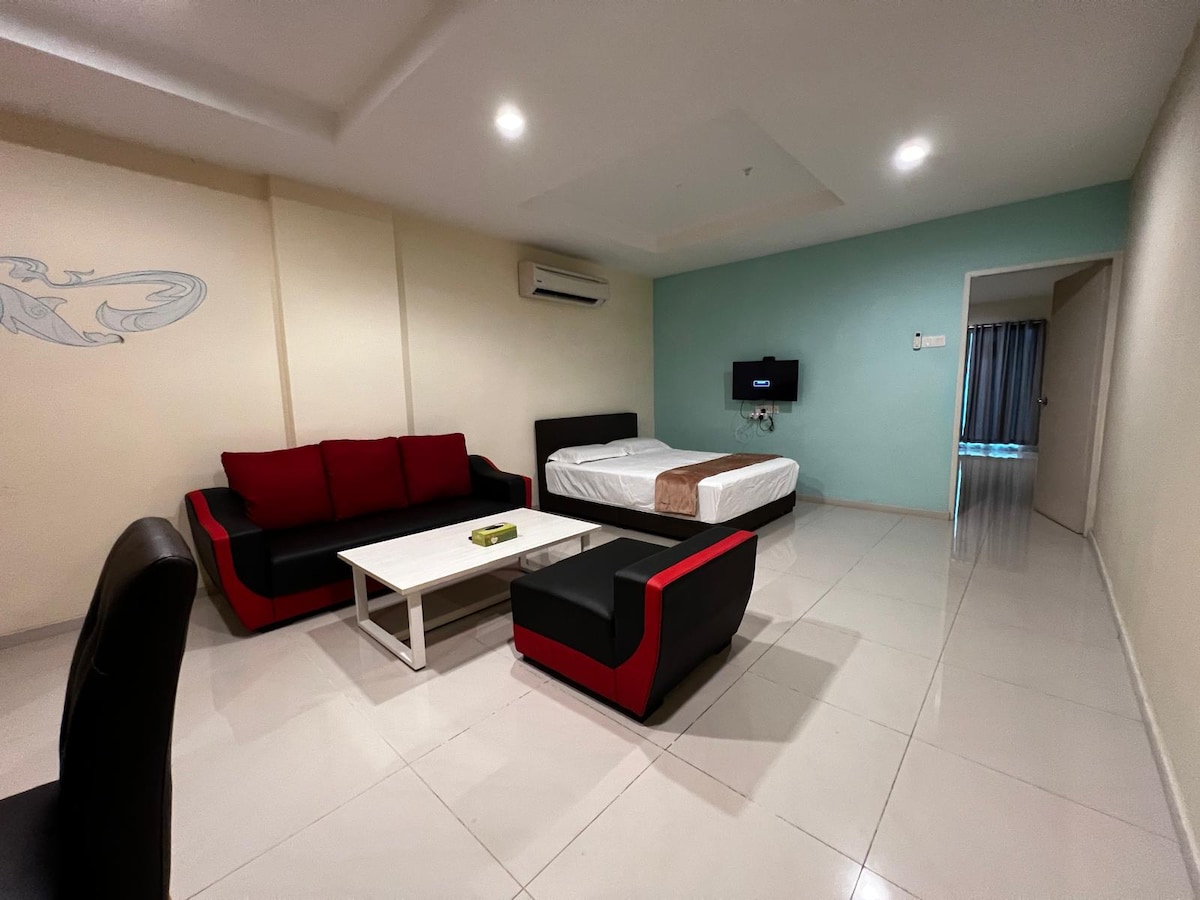 A cozy living space features a black and red seating arrangement with a coffee table at its center. A spacious bed is positioned against a light-colored wall, while a wall-mounted television is visible. Natural light enters through the partially open curtains, illuminating the room's modern design.