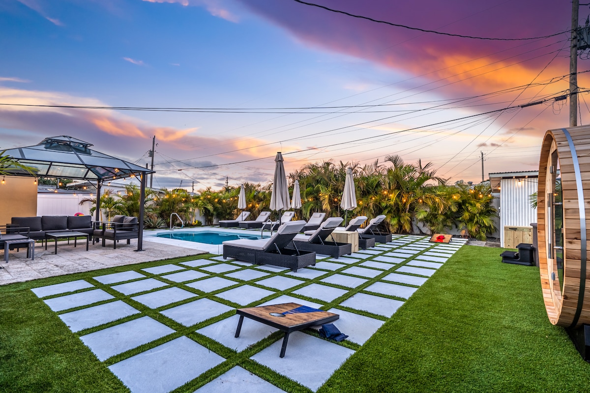 A vibrant outdoor area is depicted, featuring a sparkling pool surrounded by lounge chairs under a serene sunset sky. A traditional wood barrel sauna sits nearby, with a covered gazebo providing shade over additional seating. The lush greenery enhances the relaxing atmosphere.