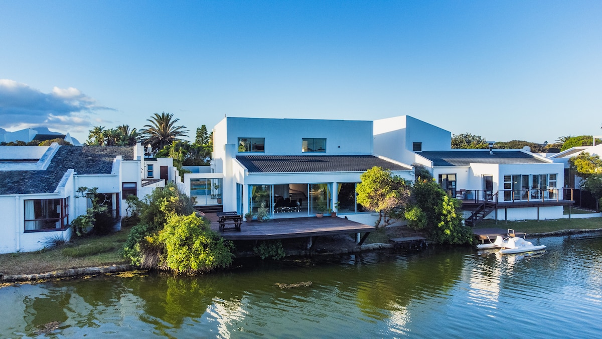 A modern waterfront house is shown, featuring a spacious deck that overlooks tranquil waters. The structure displays clean lines and large windows, allowing natural light to fill the interior. Surrounding greenery complements the serene setting along the canal.