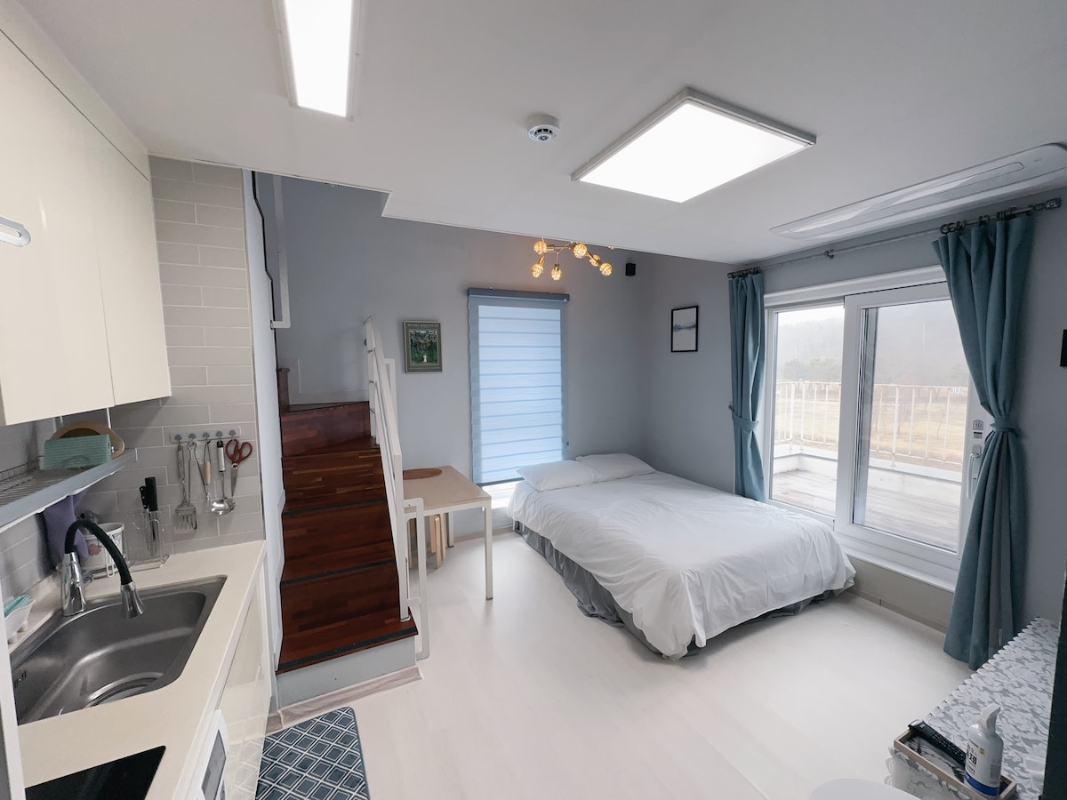 A modern studio apartment features a spacious bed with crisp white linens. Natural light enters through large windows, complemented by soft blue curtains. A compact kitchen area includes essential amenities, while a small table is positioned nearby. A staircase leads to additional sleeping space.