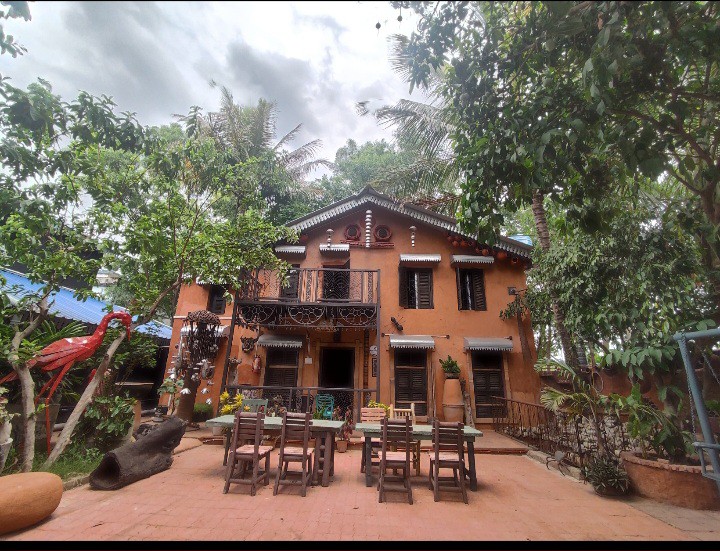 The earthy exterior of a two-story mud house is surrounded by lush greenery, showcasing a blend of artistic features. A spacious outdoor dining area with wooden furniture is positioned in front, while vibrant accents, including a decorative sculpture, enhance the inviting ambiance.