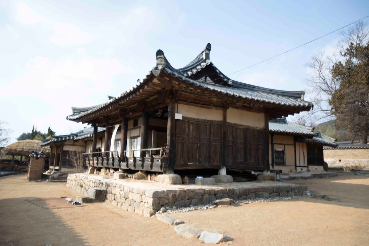 A traditional Korean hanok features wooden beams and a tiled roof, set on a raised stone foundation. The structure's wide verandah invites natural light, while the surrounding earthy landscape highlights the serene, historical ambiance of the property.