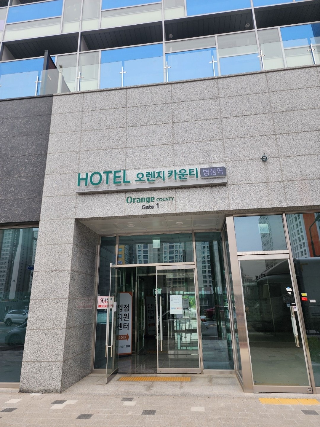The entrance to the property displays a modern facade, featuring large glass doors that lead into the building. A sign mounted above the doorway indicates 'HOTEL 오렌지카운티' and 'Orange COUNTY', welcoming guests to the space.