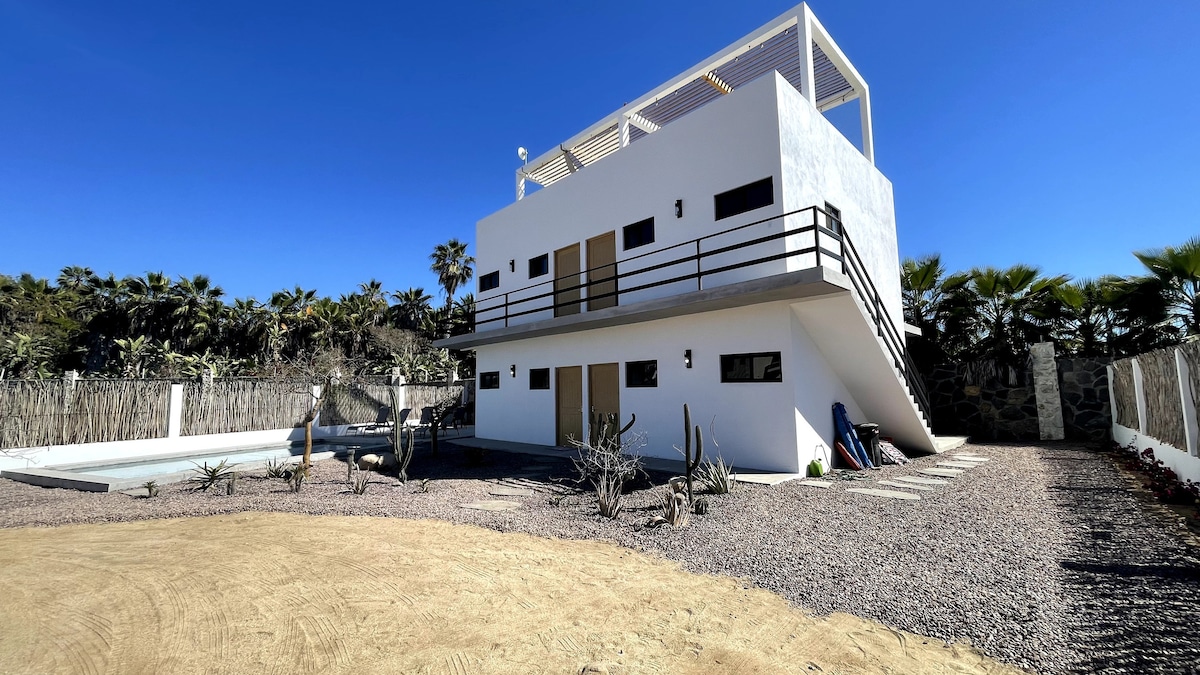 A modern two-story building is surrounded by desert landscaping and palm trees. It features a minimalist exterior with large windows and a balcony on the upper floor. A pathway leads to the entrance, and a swimming pool is located nearby, inviting relaxation.