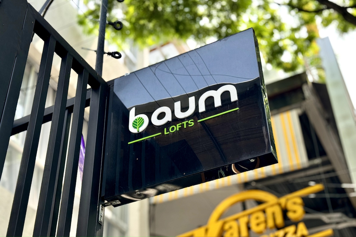 The image displays the exterior sign of BAUM Lofts, featuring a modern design with the name 'baum' in white letters on a black background, accompanied by a green leaf logo. The sign is positioned above the entrance, surrounded by trees and buildings.