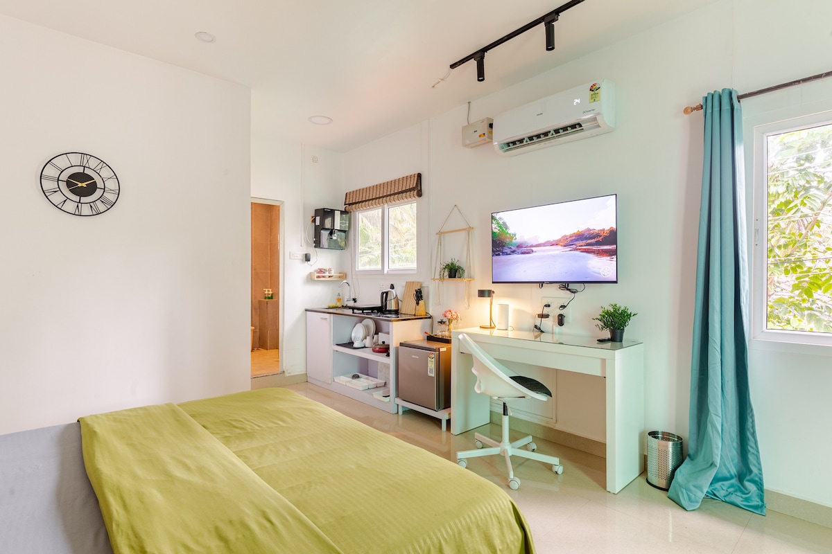 The studio features a spacious bed covered with a light green blanket, complemented by a sleek desk and chair. A flat-screen television is mounted on the wall, while a kitchenette with essential appliances is visible. Natural light enters through a nearby window, enhancing the room's bright ambiance.