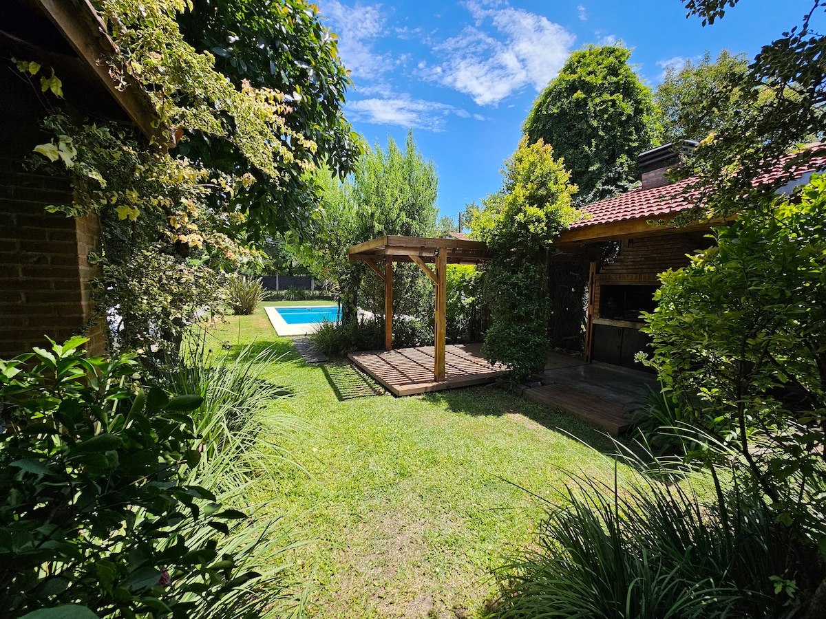A lush garden area features a wooden pergola surrounded by greenery. In the background, a swimming pool glimmers under the sunlight, framed by vibrant foliage and trees that provide natural shade.
