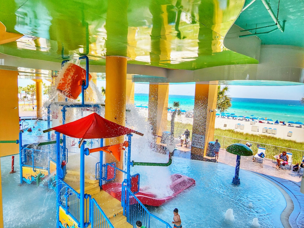 A vibrant aqua park is displayed with colorful slides and playful water features, capturing the joyful atmosphere of families enjoying the space. In the background, views of the beach and ocean are visible, enhancing the inviting setting for recreation and relaxation.
