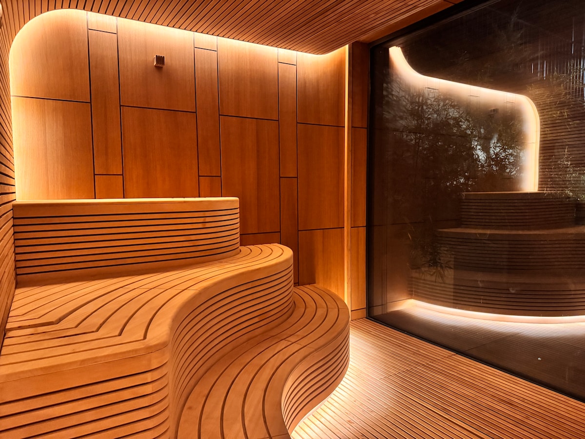 A modern sauna space is highlighted by warm wooden benches, arranged in a tiered style. Soft lighting enhances the natural wood textures, while a large glass window offers views of a serene outdoor area.