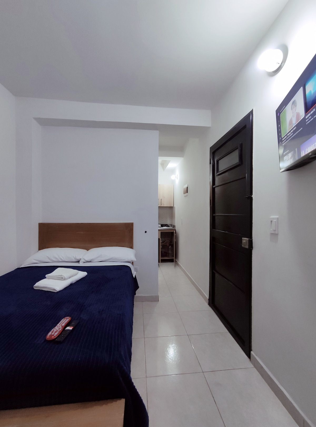 A cozy bedroom is equipped with a comfortable semi-double bed covered in a dark blue blanket. A small television is mounted on the wall, and a doorway leads to a kitchenette area. Natural light is provided through a light ceiling fixture.