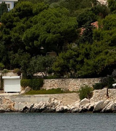The image depicts a coastal scene featuring a stone wall adorned with greenery, providing a natural border along the shoreline. A garage with a closed door is visible, nestled among the surrounding trees and plants, creating a serene and private atmosphere.