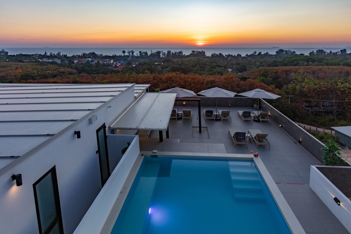 A serene view of the sunset over the horizon can be seen from the pool area, which features lounge chairs and umbrellas. The modern design of the building is complemented by the tranquil blue water of the pool, creating a relaxing atmosphere.