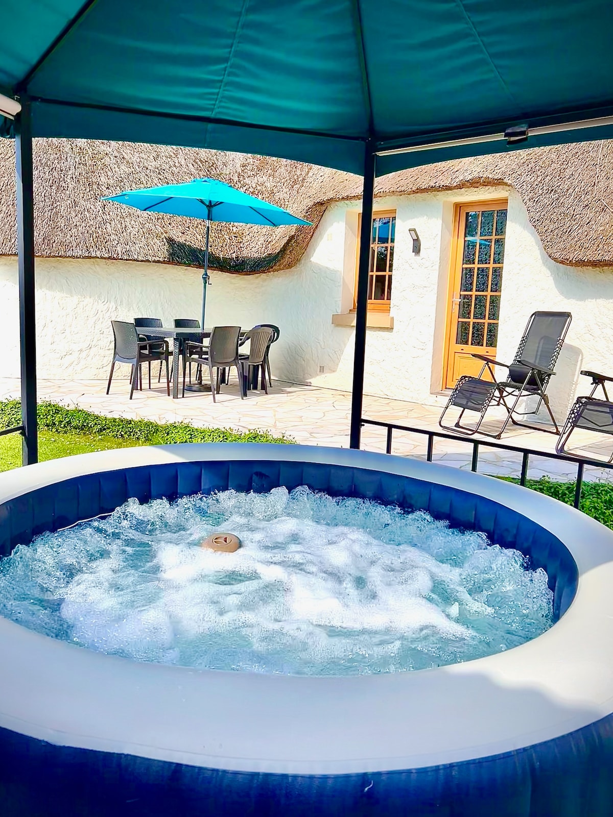 An outdoor spa is positioned under a shaded canopy, surrounded by green grass. A patio set with chairs and a table is visible in the background, accompanied by a vibrant blue umbrella. The walls of a charming house complete the serene setting.
