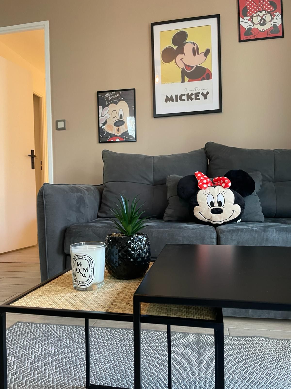 The living area features a gray sofa adorned with a Minnie Mouse cushion. A low black coffee table sits in front of the sofa, accompanied by a decorative plant and a candle. Framed Mickey Mouse prints decorate the beige walls, adding a playful touch.