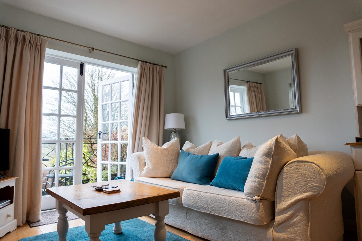 A comfortable living area is featured, with a light-colored couch adorned with decorative pillows. A wooden coffee table sits in front of the couch, and large glass doors lead to a balcony, allowing natural light to brighten the space. A mirror reflects the cozy environment.