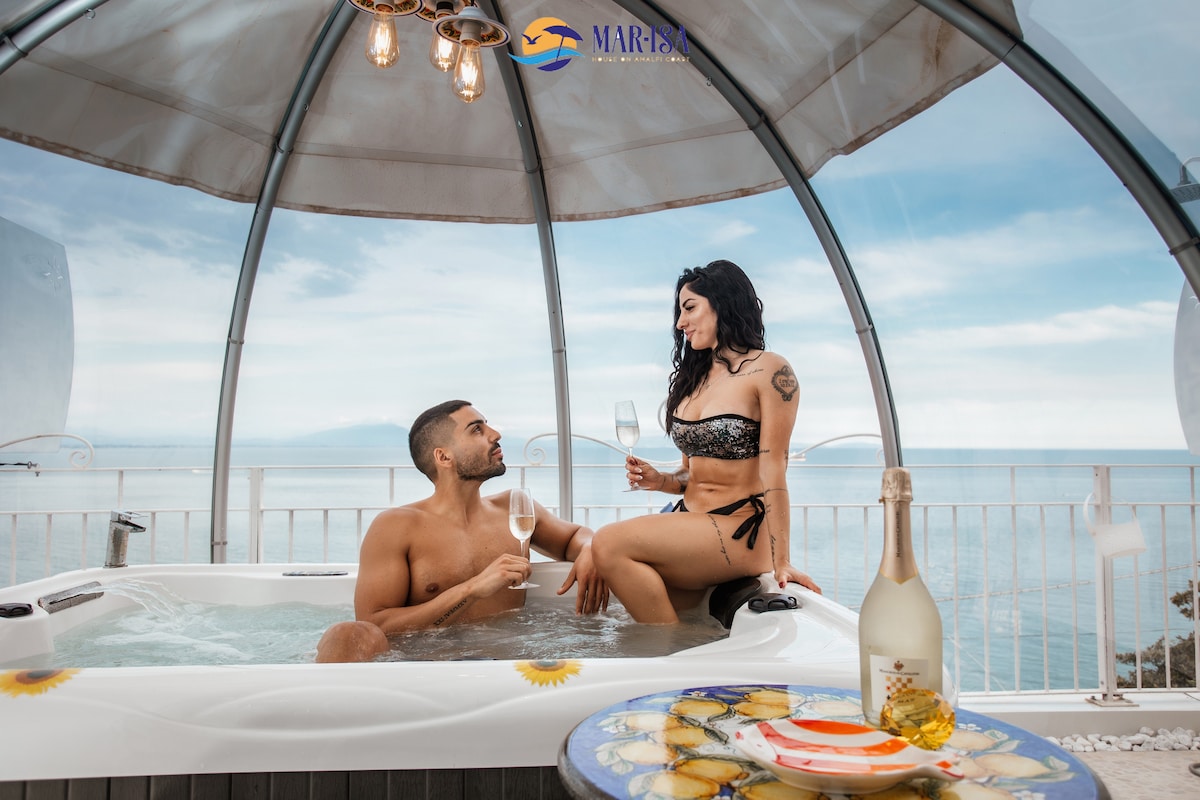 A bubble-shaped structure features an outdoor hot tub with two guests enjoying the setting. The sea view is visible in the background, complemented by soft natural lighting. A bottle of champagne and fluted glasses are placed nearby, enhancing the ambiance of the relaxed environment.