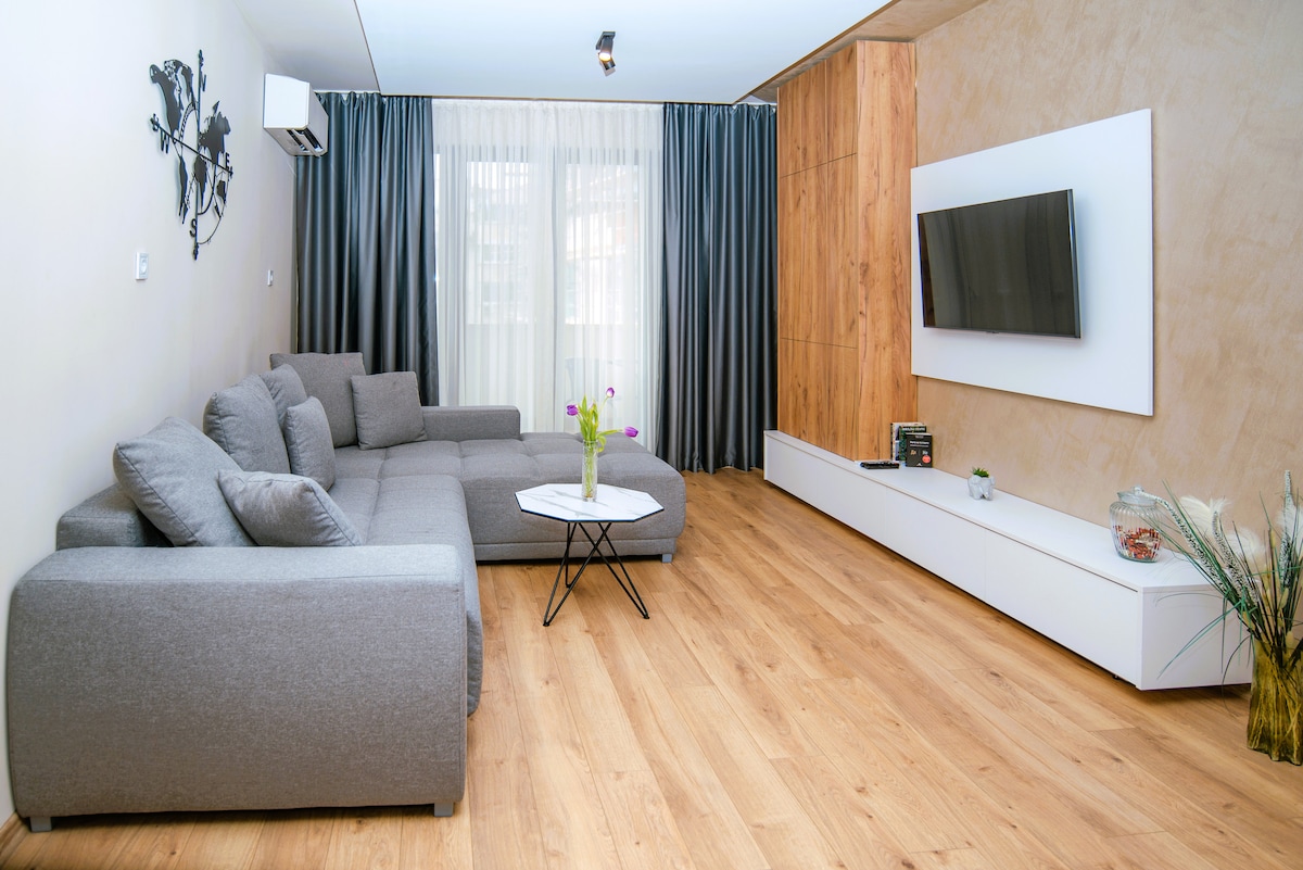 A spacious living area features a modern gray sectional sofa arranged around a white coffee table. Light wood flooring complements the room, illuminated by natural light through large windows dressed with sheer and dark curtains. A mounted television and decorative accents add to the design.