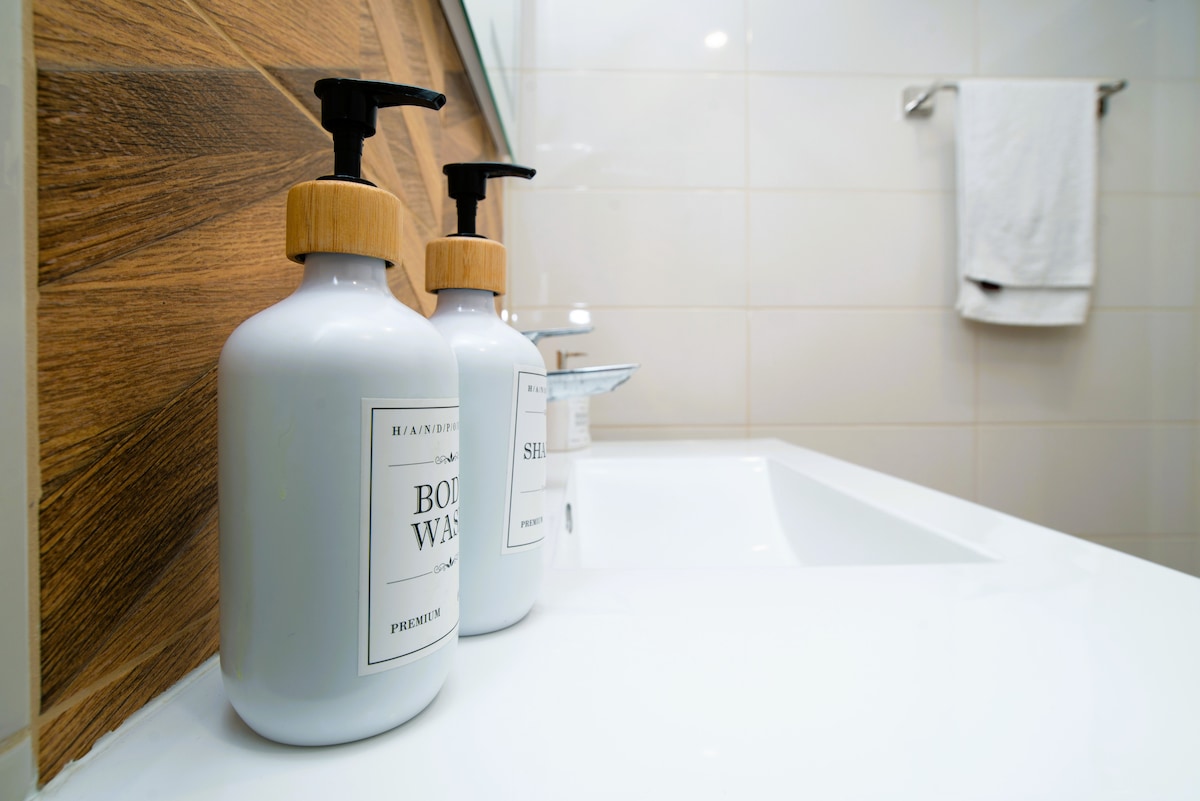 A bathroom vanity displays two elegant soap dispensers with wooden accents, positioned beside a sleek, white sink. Soft beige tiles create a warm atmosphere, complemented by a neatly hung white towel in the background.