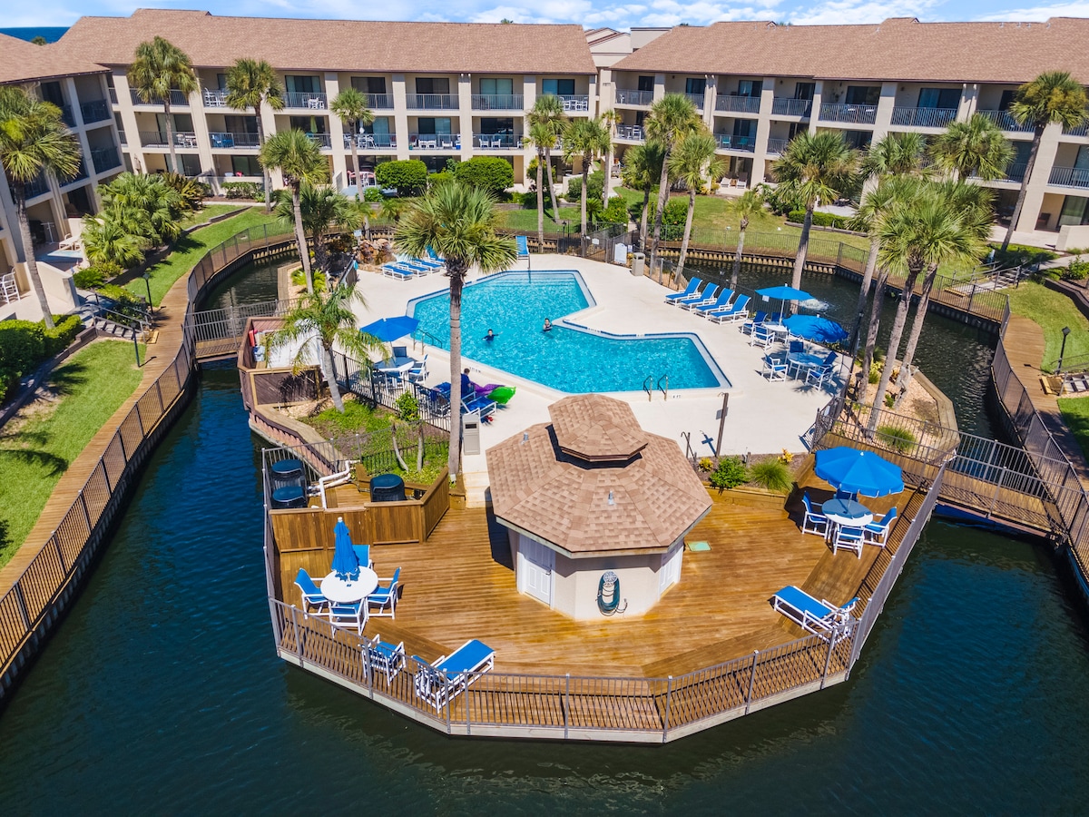 An aerial view captures a spacious pool area surrounded by well-maintained landscaping and palm trees. Sun loungers and umbrellas are arranged around the pool, which is shaped like a lagoon. Multiple pathways lead to the pool, enhancing accessibility for all guests.