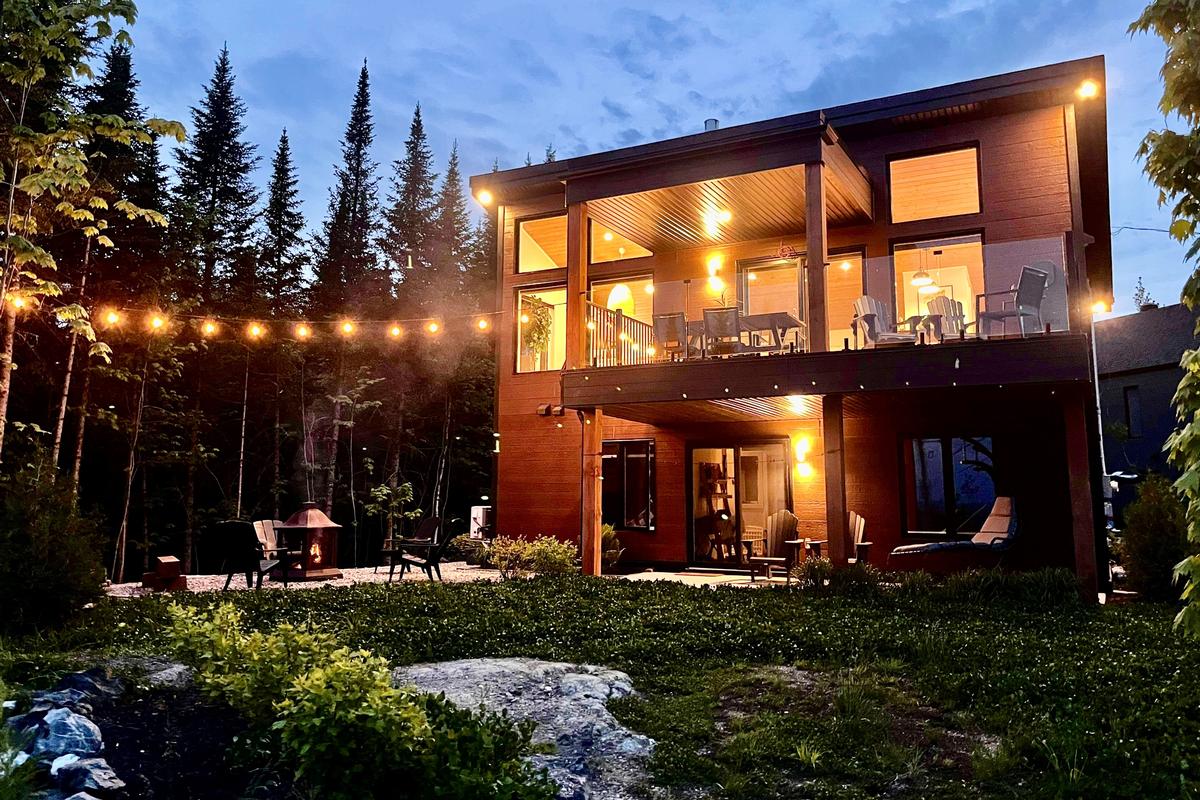 The chalet's exterior is illuminated at dusk, showcasing a modern design with large windows and a spacious terrace. Strings of lights create a warm ambiance, while surrounding greenery enhances the natural setting. Outdoor seating areas are visible, inviting relaxation in the serene environment.