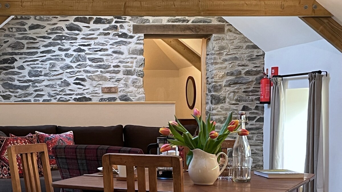 An open-plan living space features a cozy seating area with dark cushions and vibrant throw pillows across from a dining table. Fresh tulips are displayed in a vase, while warm stone walls provide a rustic backdrop. Natural light enters through nearby windows.