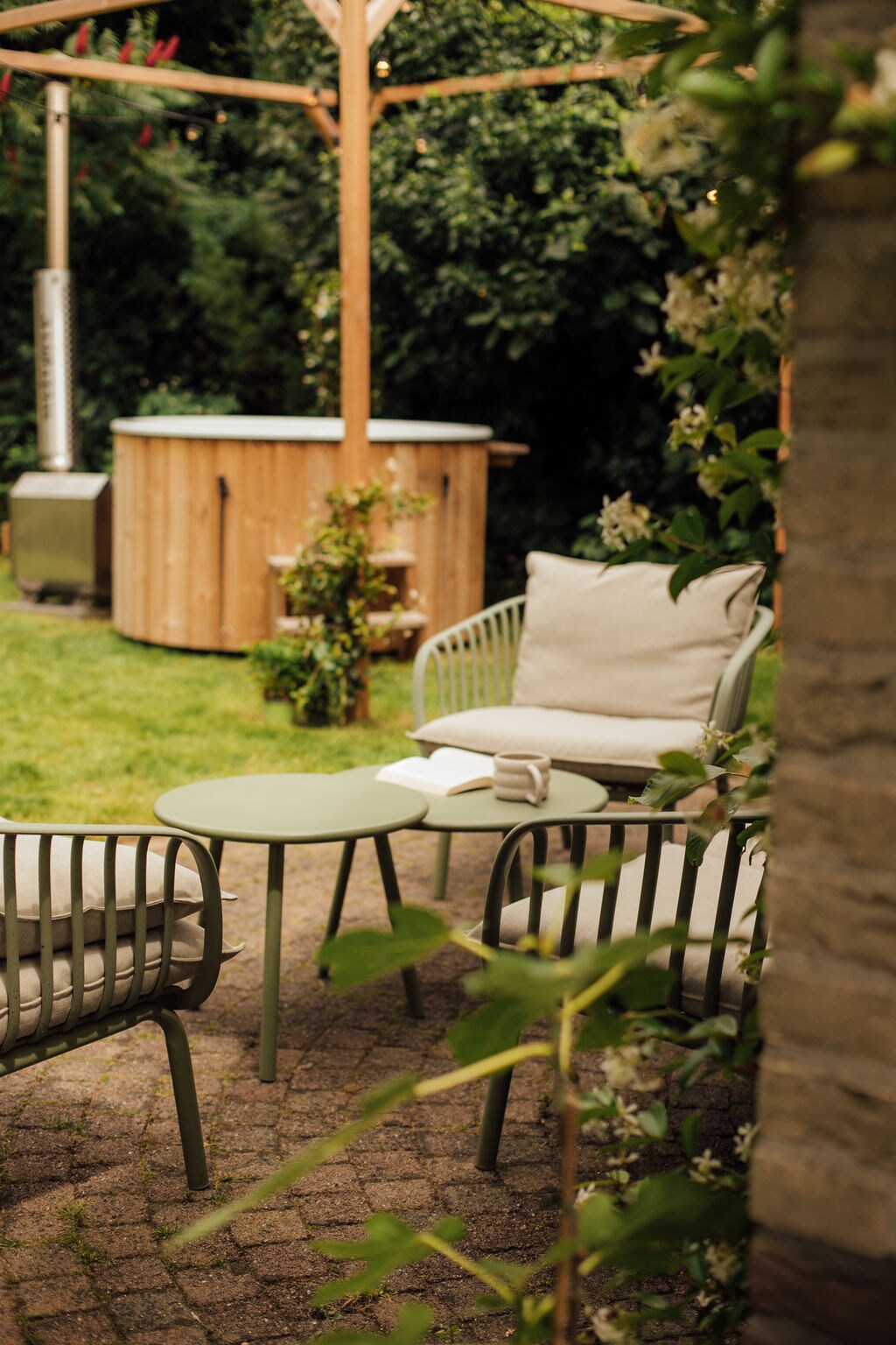 A cozy outdoor seating area is featured, consisting of two armchairs and a circular table set on a stone patio. A hot tub is located in the background, surrounded by green foliage, contributing to the serene garden atmosphere.