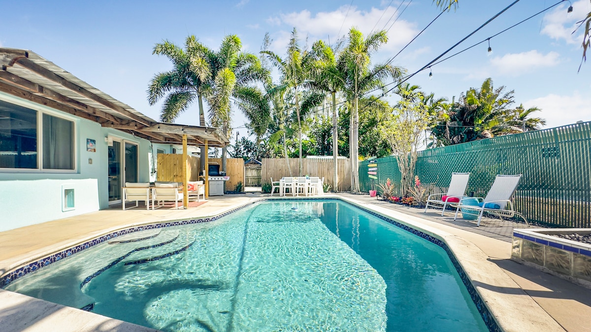 A private heated pool is surrounded by palm trees and a spacious patio area, featuring lounge chairs and a nearby outdoor dining set. Sliding glass doors provide access to an inviting living space, blending indoor and outdoor areas harmoniously.