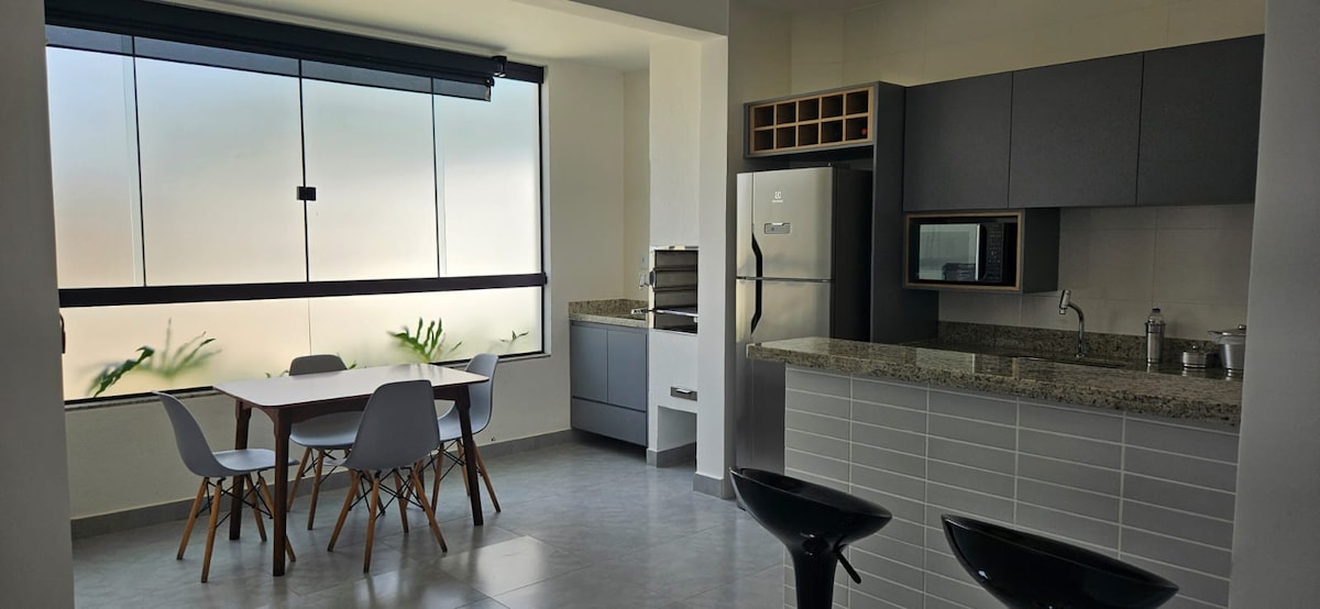 A modern kitchen and dining area features a granite countertop and stainless-steel appliances, including a refrigerator and microwave. A table with four chairs is set by a large window adorned with plants, providing natural light to the space.