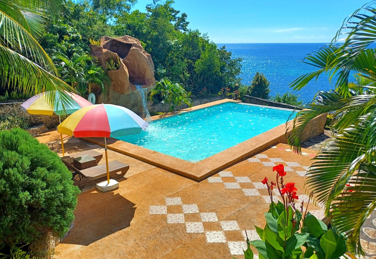 A freshwater pool is surrounded by lush greenery and colorful umbrellas, offering a refreshing escape. A waterfall feature adds a tranquil sound, while the ocean view creates a serene backdrop for relaxation. The pool area includes a spacious patio laid with decorative tiles.