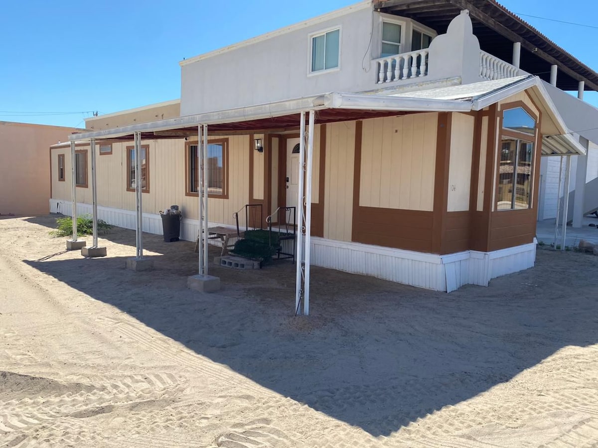 The exterior of the house features a covered porch supported by two columns, providing shade to the entrance. Windows allow natural light to fill the interior. A sandy area surrounds the structure, emphasizing its outdoor space. The roofline displays a decorative ledge.