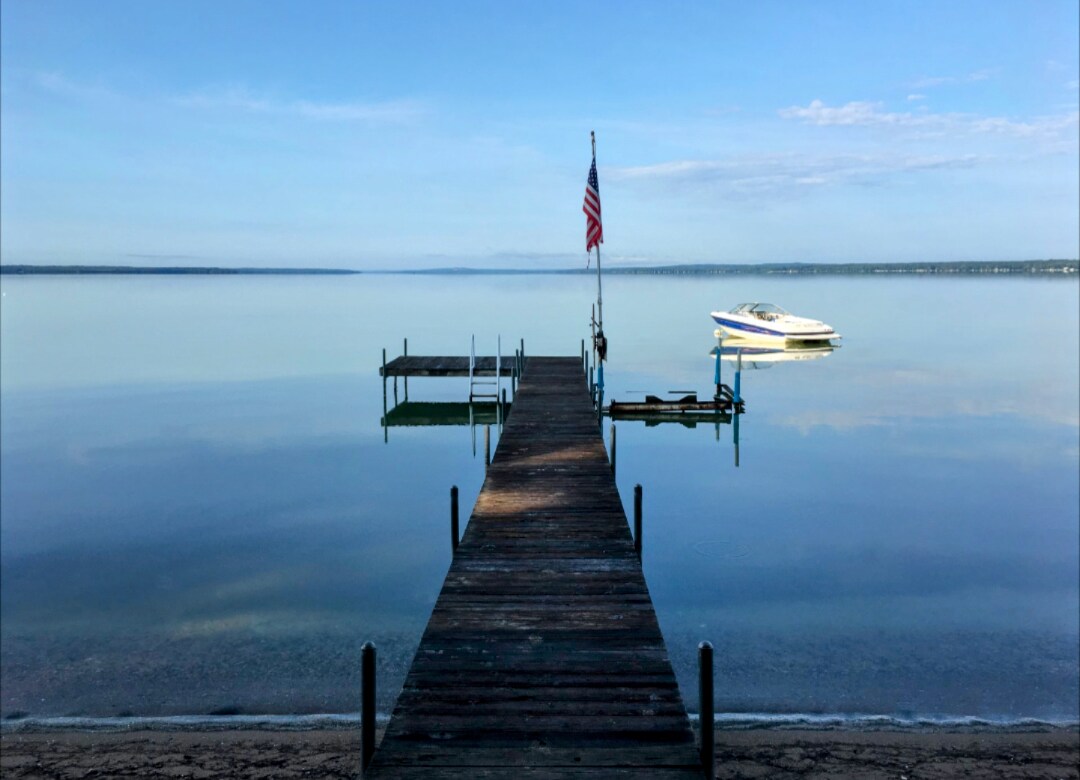 A wooden dock extends over a calm lake, with an American flag prominently displayed. A boat is anchored nearby, and the serene water reflects the sky, creating a peaceful atmosphere. The sandy shore is visible in the foreground, leading to the dock.