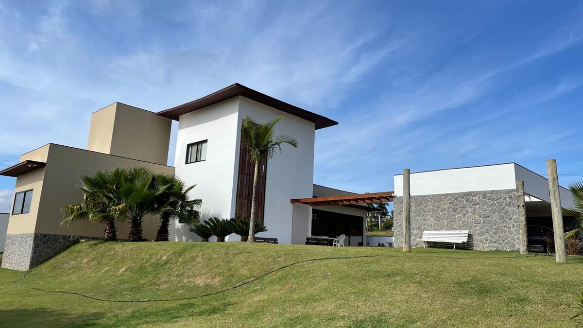 A modern multi-level house is set amidst lush greenery, with large windows and a blend of stone and white walls. The front yard features a well-manicured lawn, accentuated by palm trees and outdoor seating under the shaded roof.