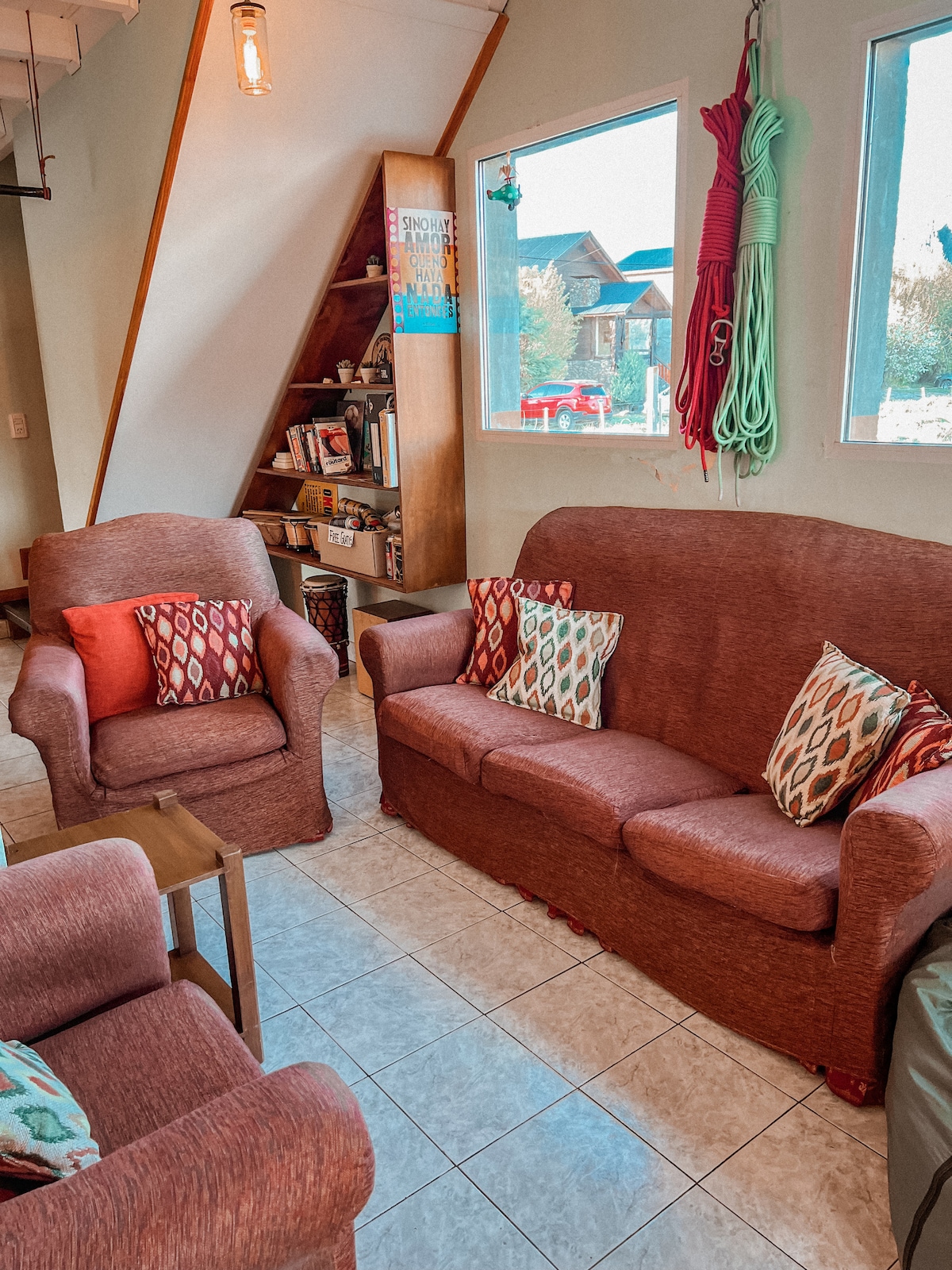 A comfortable common area features two inviting sofas and two armchairs, all adorned with patterned cushions. Natural light filters through large windows, creating a bright atmosphere. A bookshelf displays various items, contributing to the cozy, relaxed environment.