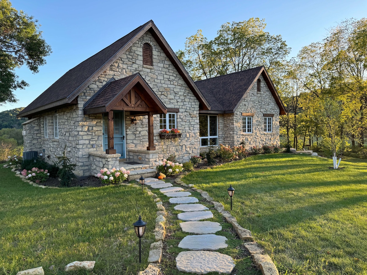 A charming stone cottage sits amidst lush greenery, featuring a well-defined pathway leading to an inviting front entrance. Softly lit lanterns line the walkway, while flowerbeds enhance the cottage's exterior. Trees provide shade, complementing the serene landscape under a clear blue sky.
