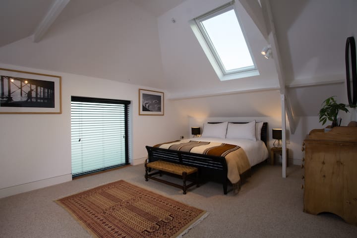 เตียงขนาดคิงไซส์ขนาดใหญ่โต๊ะข้างและโคมไฟ คานโครงสร้างเดิมที่ทาสีขาว หีบลิ้นชักและกระจกโบราณ หน้าต่าง velux ขนาดใหญ่ 2 บาน (บานหนึ่งมองเห็นวิวหุบเขา) รูปถ่ายขาวดำกรอบดั้งเดิมถ่ายโดยเจ้าของที่พัก