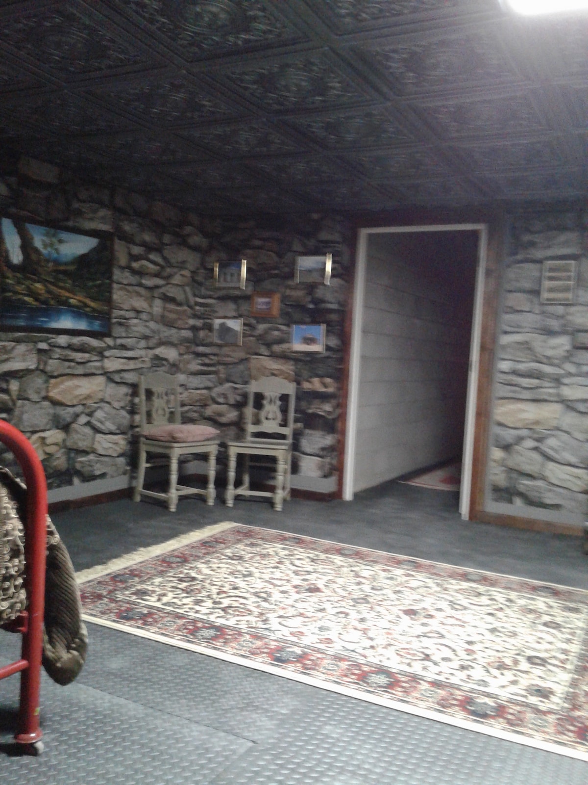 A rustic room features stone walls and a patterned ceiling. Two decorative chairs are positioned against the wall, accompanied by framed artwork. A patterned area rug covers the floor, and a doorway leads to another space.