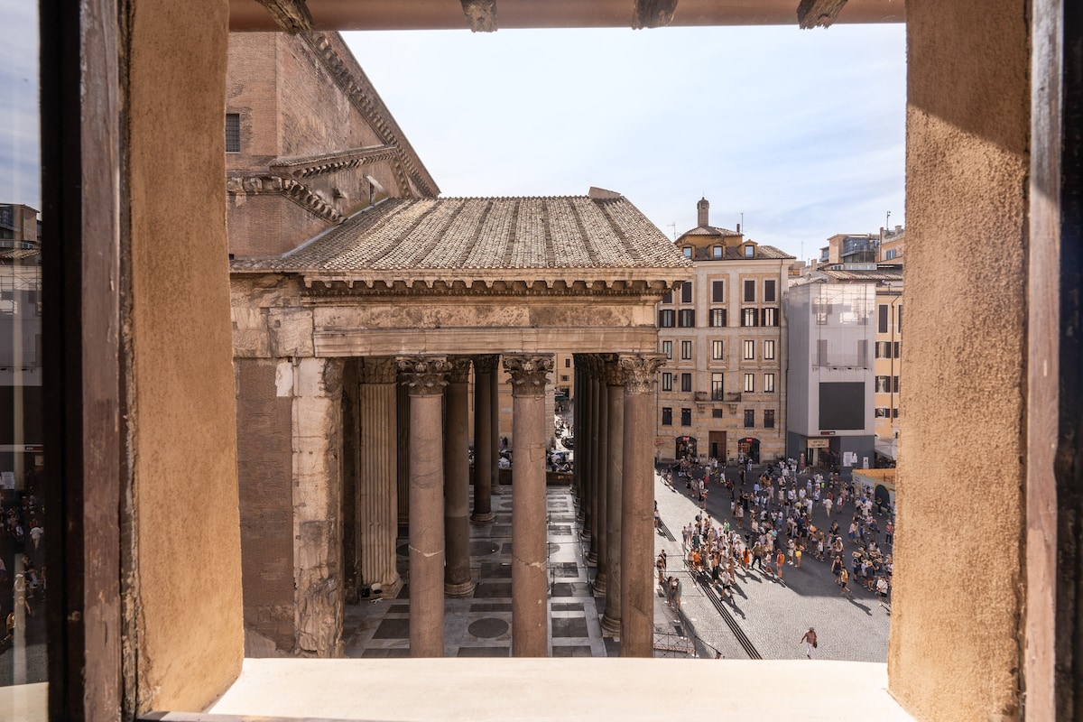 A view from a window frames the bustling Piazza del Pantheon below. Stone columns of the ancient temple are visible, alongside crowds enjoying the square. The warm sunlight illuminates the vibrant activity outside, revealing architectural details of historical buildings surrounding the plaza.