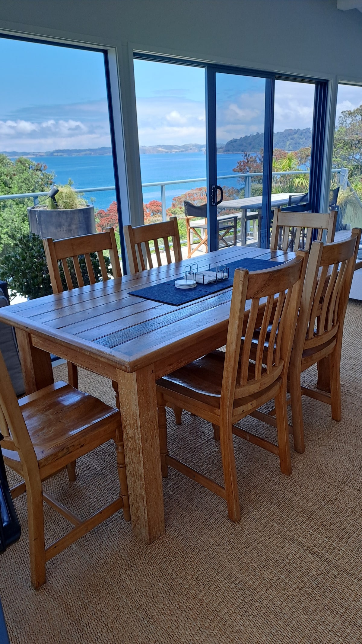 A wooden dining table surrounded by six matching chairs is positioned beside large glass doors, offering a view of Omaha Bay. Natural light fills the space, and a textured rug is laid beneath the table, enhancing the serene atmosphere.