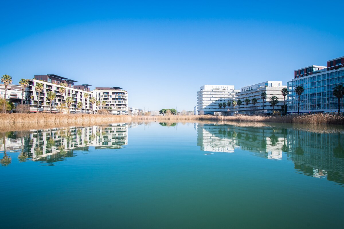 A serene waterway reflects modern apartment buildings and palm trees along its banks. The clear blue sky enhances the peaceful atmosphere, creating a harmonious connection between the urban landscape and nature.