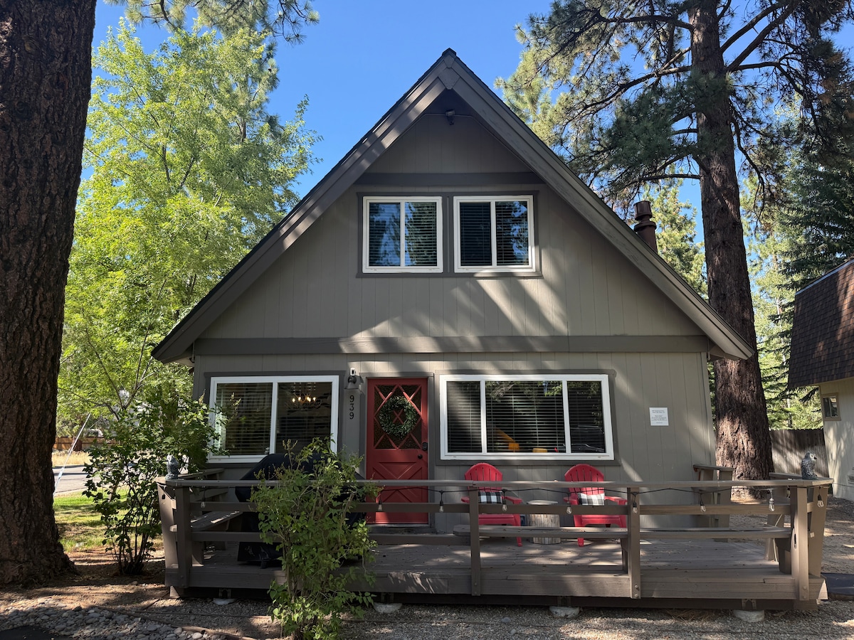 The exterior of the Tanglewood Cabin is presented with a sloped roof and a welcoming red door. Surrounded by tall trees, a spacious front deck features two bright red chairs and a low railing, inviting relaxation in a natural setting.