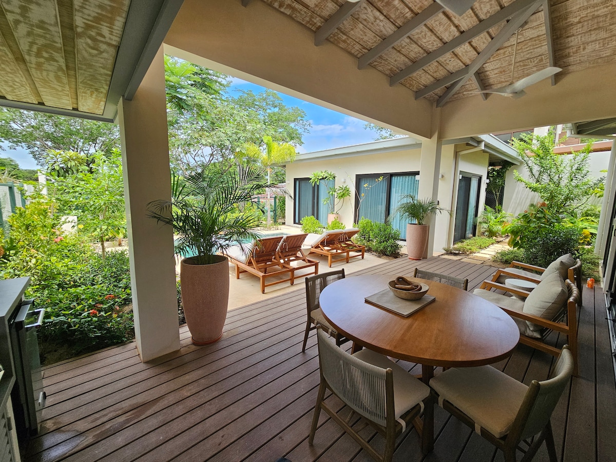 A covered outdoor dining area features a round table surrounded by four chairs. Sun loungers are positioned near a lush garden, which includes potted plants. Large glass doors provide views of the pool area, with tropical foliage enhancing the serene environment.