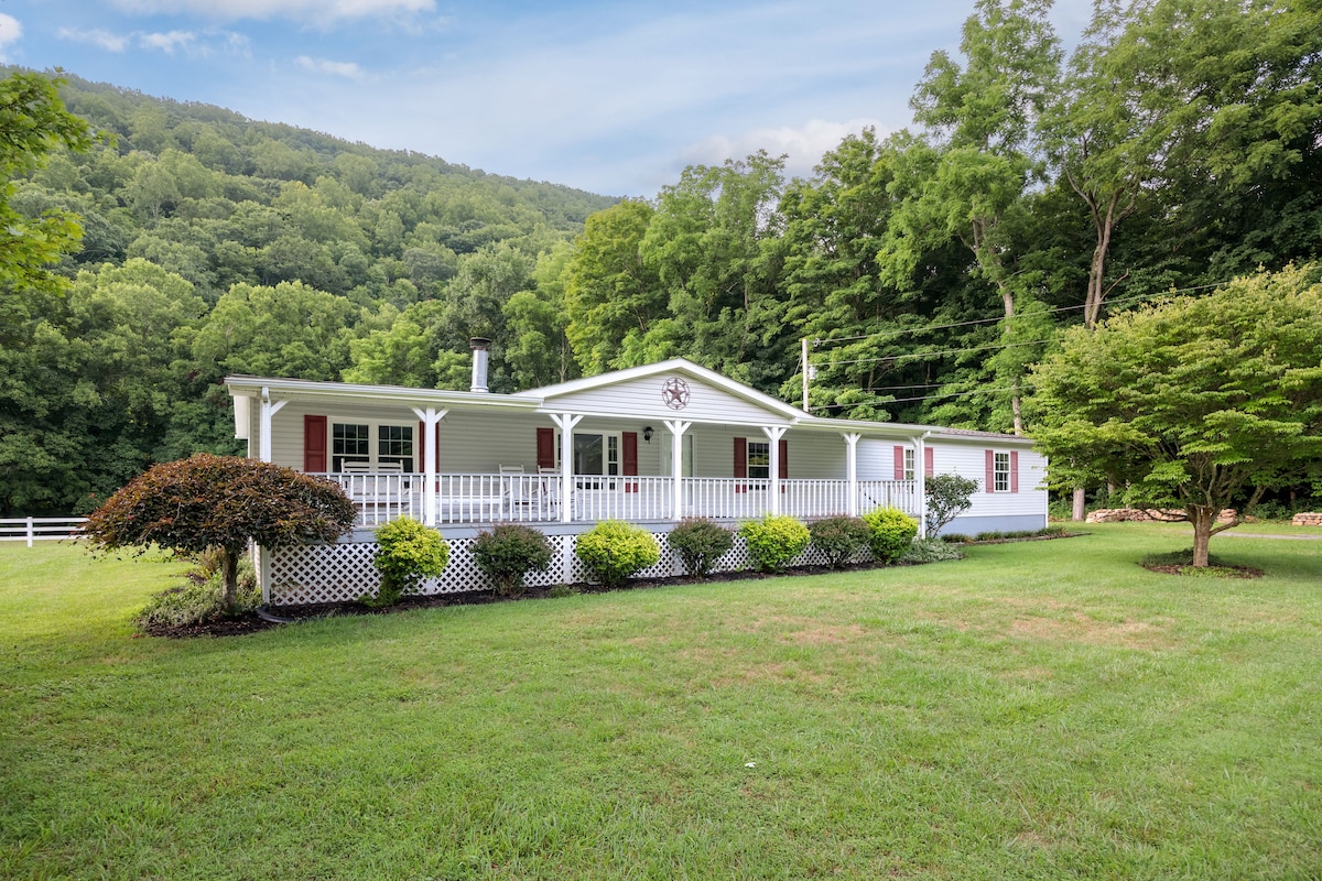 A charming, one-story house is set against a backdrop of lush green hills. The home features a welcoming front porch with a white railing, surrounded by well-maintained shrubs and a manicured lawn, inviting guests to relax and enjoy the serene environment.