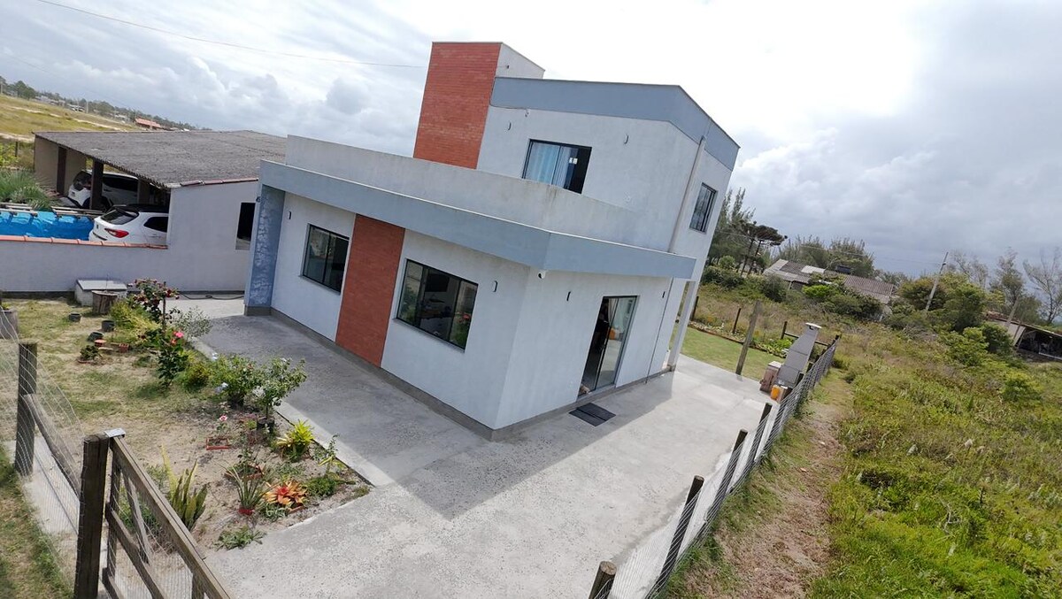 The exterior of the house showcases a modern two-story design with a combination of light-colored walls and red accents. A concrete path leads to the entrance, bordered by landscaped gardens. The property is situated in a tranquil setting with open views of the surrounding area.