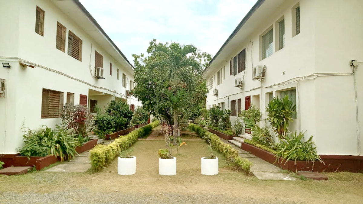 An outdoor area is showcased, featuring two parallel buildings with white exteriors. Lush greenery surrounds a central grassy space, with well-maintained flower beds and planters. Air conditioning units are visible on the buildings, enhancing the comfort of the environment.