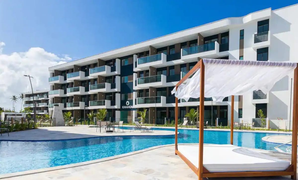 A spacious outdoor area features a large swimming pool surrounded by lush greenery. A shaded lounging cabana is positioned near the water. The modern condominium building stands in the background, showcasing its contemporary architecture and featuring multiple balconies.