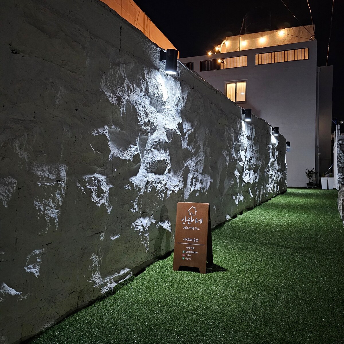 A narrow path lined with textured stone walls is illuminated by soft, focused lights, creating a serene atmosphere at night. Artificial grass covers the ground, enhancing the peaceful environment. A sign marking the property is visible at the entrance, providing orientation for guests.